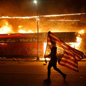 A protester carries a U.S. flag upside down, a sign of distress, next to a burning building Thursday, May 28, 2020, in Minneapolis. Protests over the death of George Floyd, a black man who died in police custody Monday, broke out in Minneapolis for a third straight night. (AP Photo/Julio Cortez)